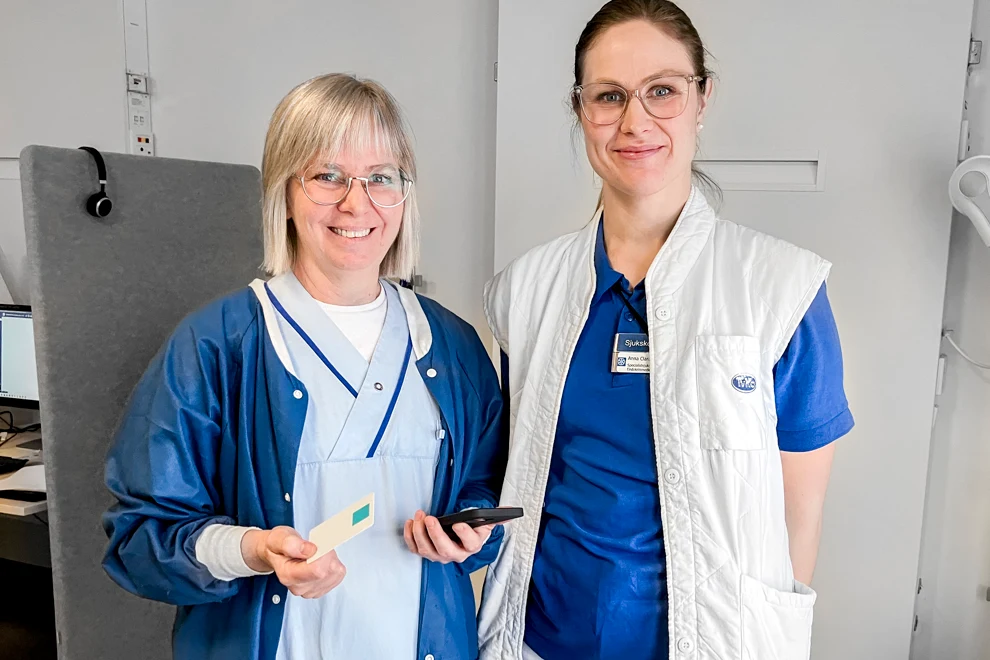 Tv kvinnor som står bredvid varandra. De bär blå vårdkläder. De ler mot kameran.