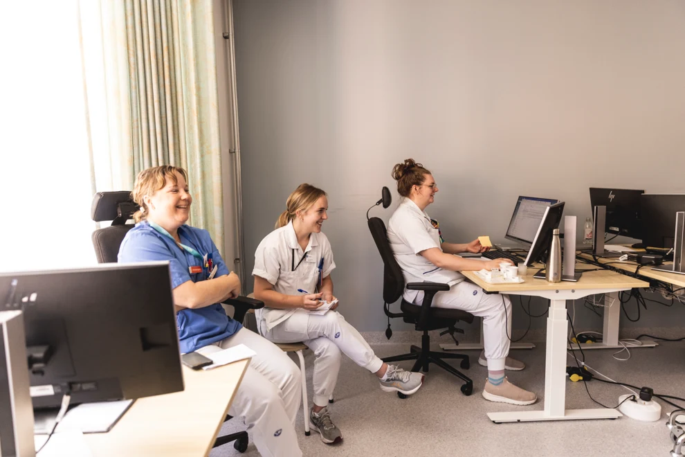 Tre kvinnor som arbetar inom vården skrattar på ett kontor med flera datorer. Väggarna är vita och golvet grått.